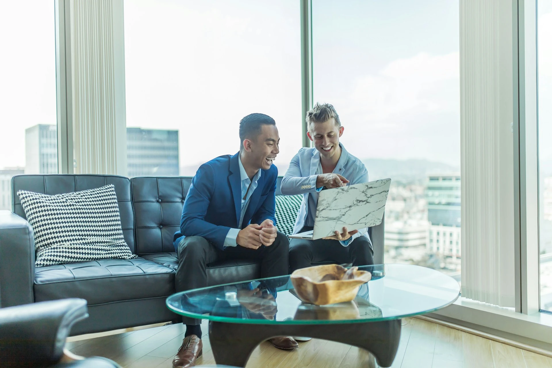 Two men in suits sit on a sofa looking at a laptop.