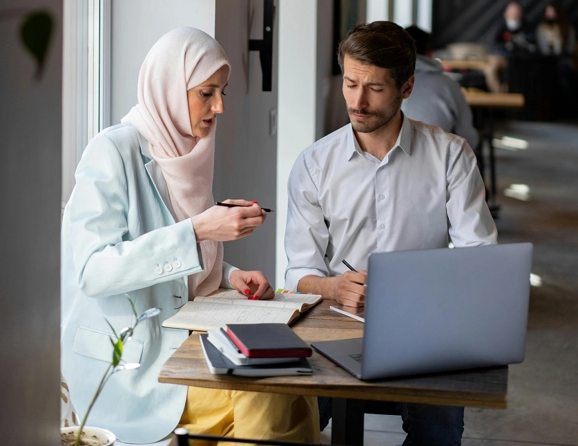 رجل وامرأة يتناقشان وأمامهما كمبيوتر محمول، وأقلام، وأوراق.