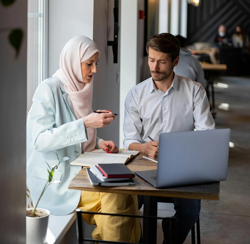 رجل وامرأة يتناقشان وأمامهما كمبيوتر محمول، وأقلام، وأوراق.
