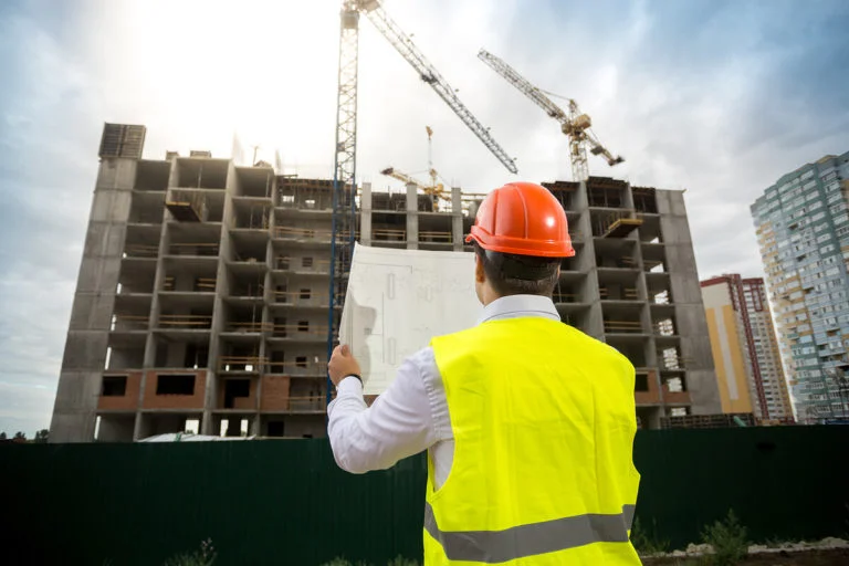 Construction worker with reflective vest and hard hat, holding a blueprint, standing in front of a high rise building under construction