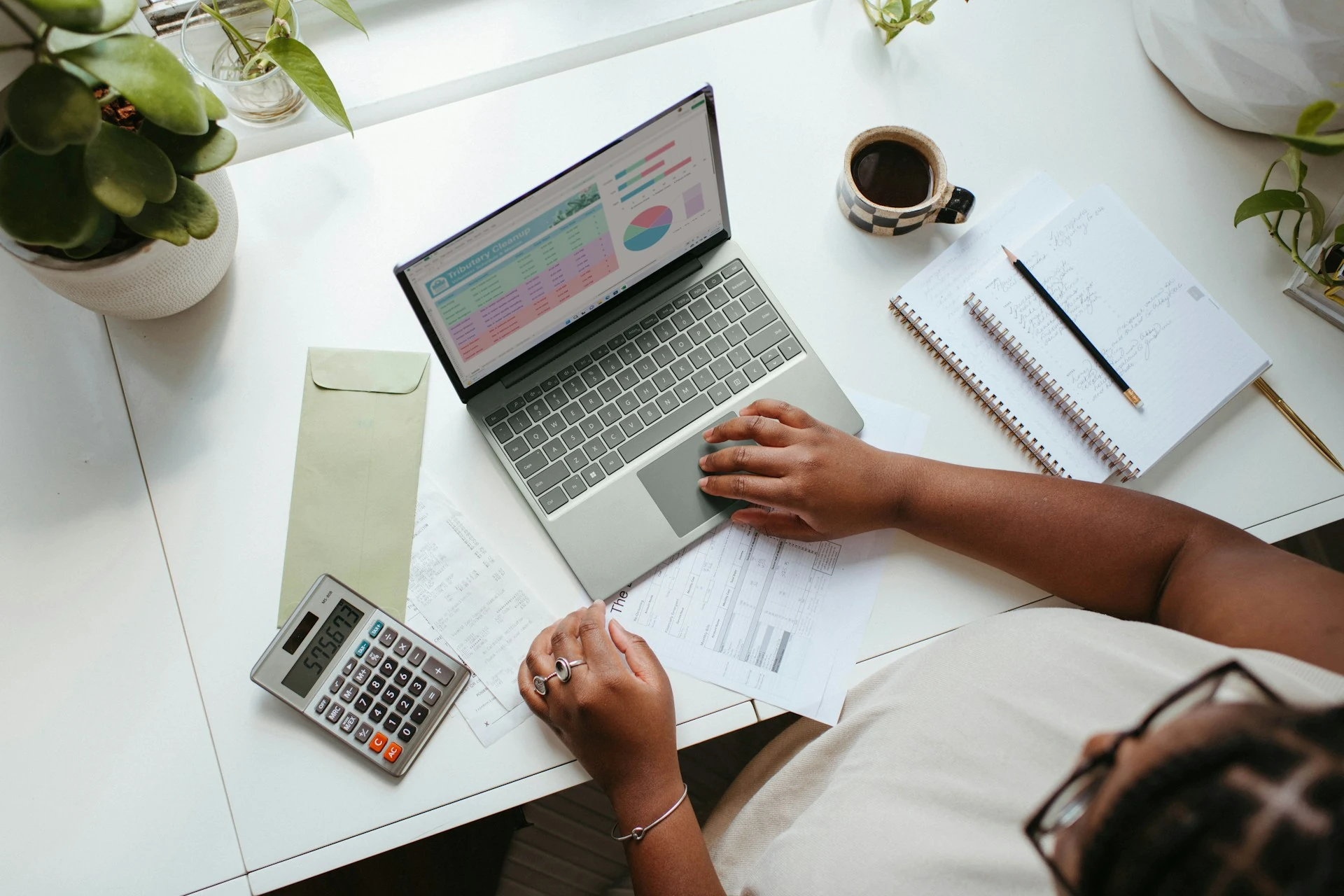 Overhead view of a woman's hands doing finances at a white desk, one hand rests on a laptop trackpad