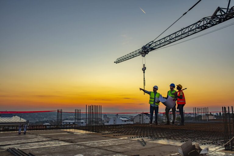 Three construction standing on top of a structure, one pointing and all three looking at something in the distance, with a crane in the background.