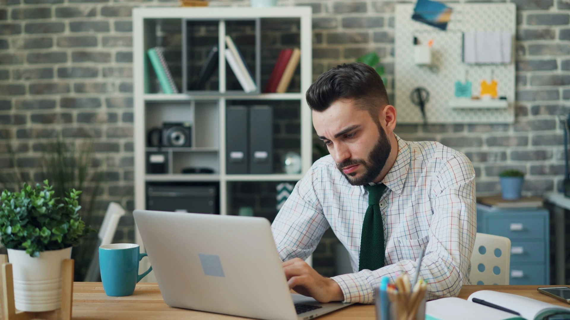 A frustrated man looking at his laptop, a blue cup of coffee next to him.