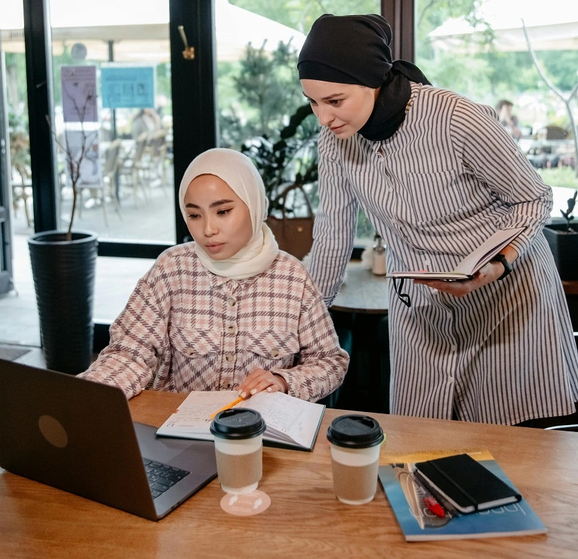 امرأتان تنظران في لابتوب وتحمل إحداهما مفكرة.