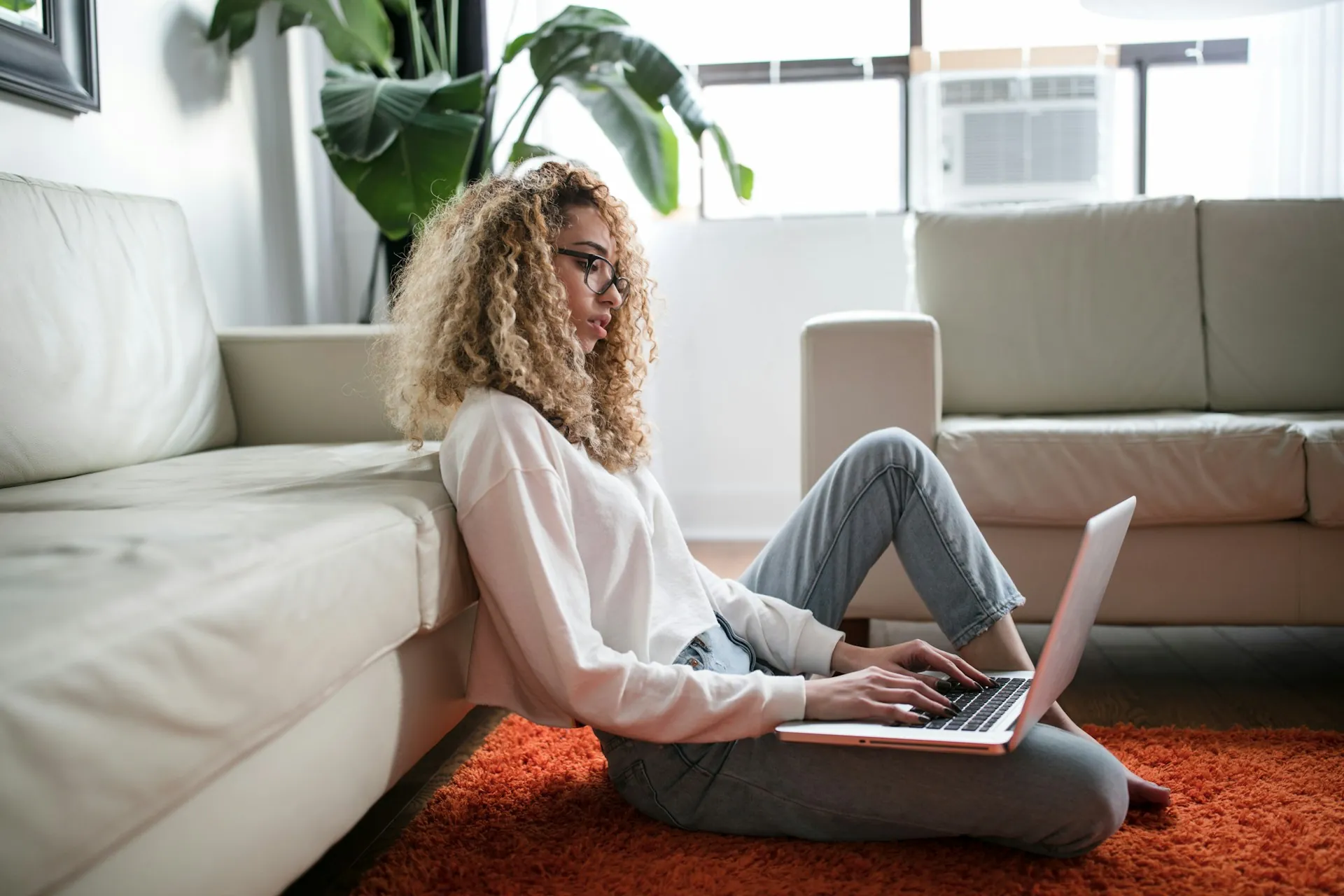 Una mujer que trabaja en su computadora portátil mientras está sentada en el piso, recostada contra un sofá.