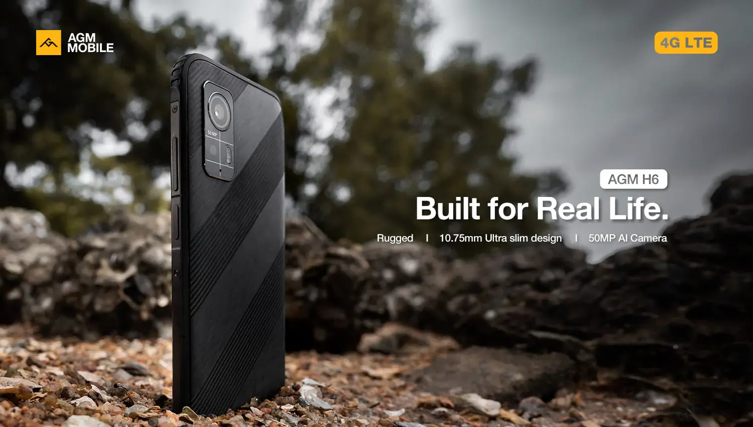 Teléfono resistente AGM H6 sobre un terreno rocoso al aire libre con árboles al fondo.