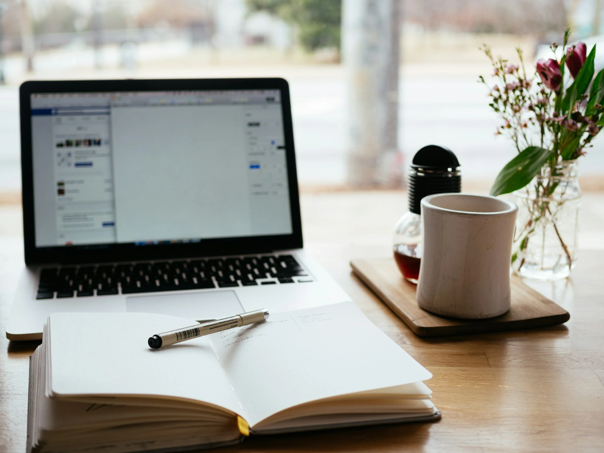 An open notebook with a pen placed on top, placed next to a laptop and cup of coffee.