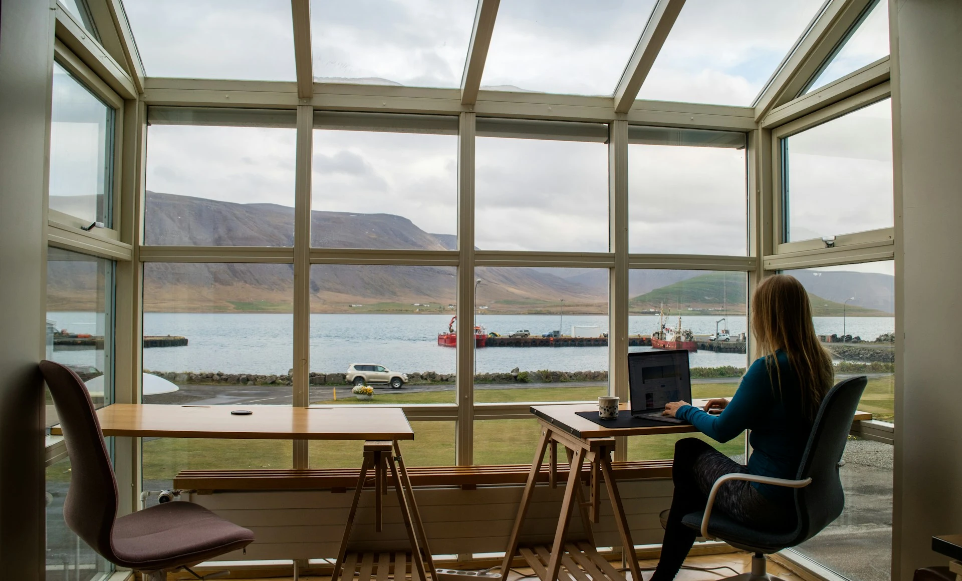A person in a blue jacket sits on a brown wooden chair near a brown wooden table during the day with the view of the. mountain outside.
