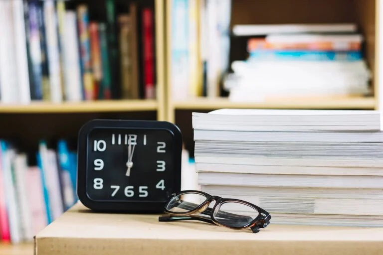 Alarm clock next to a stack of books and glasses on a desk representing work hours and time tracking.